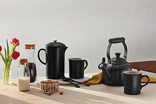 Table de cuisine avec théière noire, cafetière, tasses et fleurs.